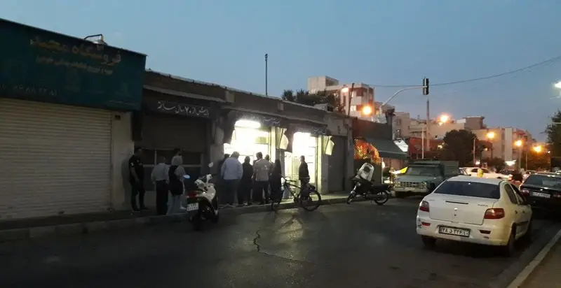 صحنه‌ای دلنشین از مغازه بستنی حاج صفر در غروب، با حضور مشتریان و خودروها در خیابان، نشان‌دهنده فعالیت پررونق این کسب و کار محبوب.