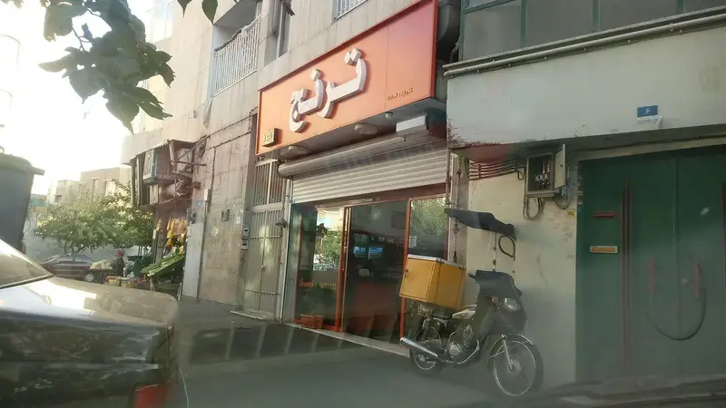 نمای بیرونی فست فود و پیتزا ترنج با تابلوی بزرگ نارنجی و موتورسیکلت تحویل غذا با باکس زرد در خیابان.