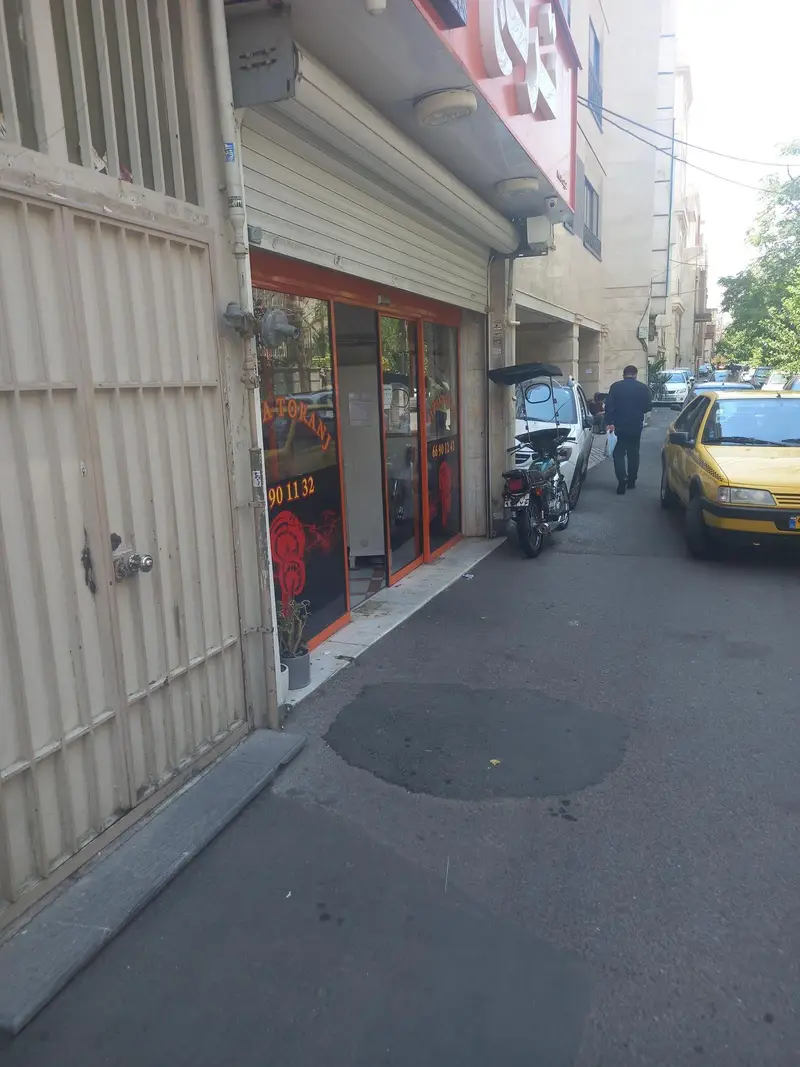 تصویر بیرونی پیتزا ترنج: ورودی شیشه‌ای، تابلوی با نام و شماره تماس، موتورسیکلت، تاکسی زرد و عابران در خیابان.