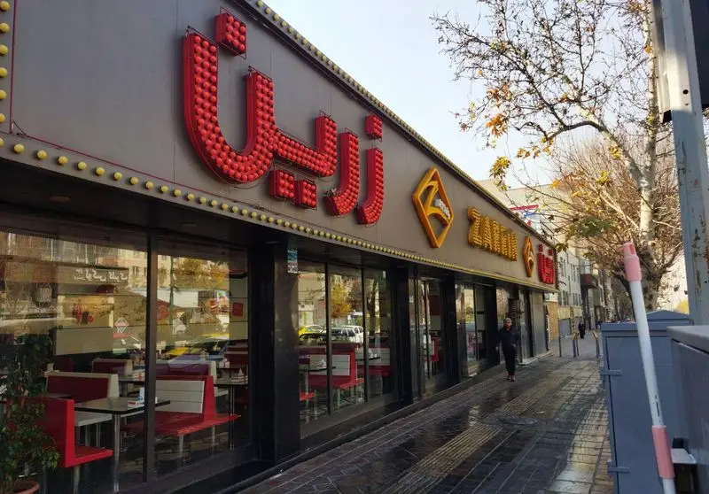 نمای بیرونی رستوران پیتزا زرین، با تابلوی بزرگ و جذاب حروف قرمز و طلایی در بالای ورودی شیشه‌ای. صندلی‌های قرمز در داخل دیده می‌شوند.