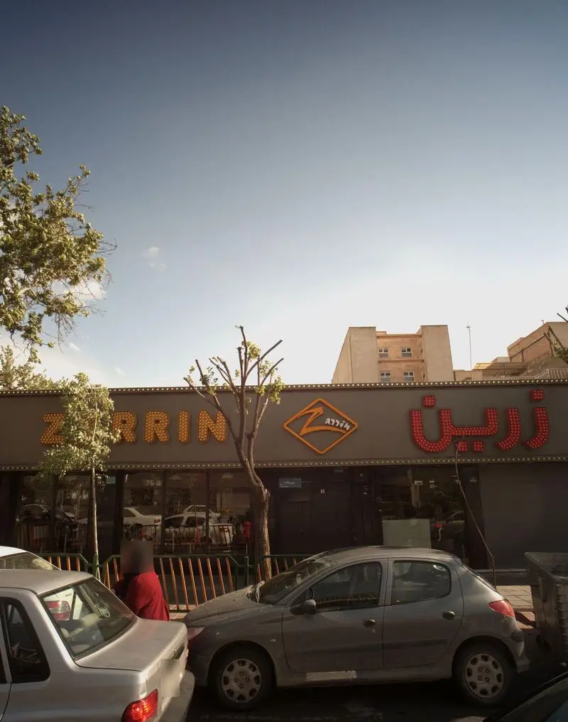 تصویر نمای بیرونی ساختمان فست فود و رستوران پیتزا زرین (Zarrin Pizza) با تابلوی درخشان، درختان و خودروها در تهران.