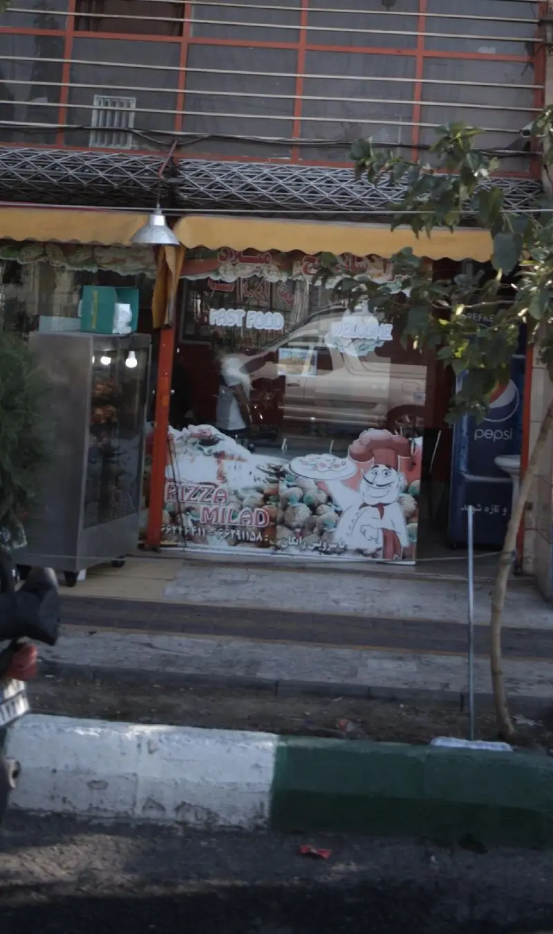 نمای خارجی فست فود پیتزا میلاد با بنر بزرگ تبلیغاتی پیتزا، نام تجاری، شماره تلفن و یخچال نوشابه پپسی در کنار خیابان.