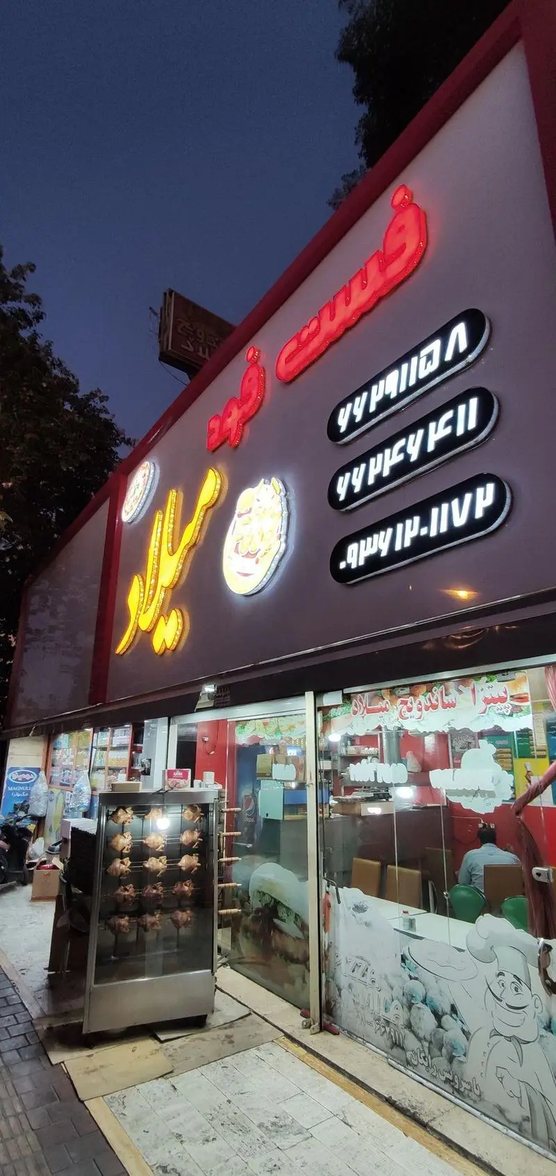 نمای بیرونی رستوران و فست فود پیتزا میلاد در شب، شامل تابلوهای درخشان، مرغ بریان و شماره تماس جهت سفارش غذا.