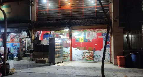 پیتزا میلاد: نمای شبانه و روشن مغازه فست فود با ویترین‌های جذاب برگر، مرغ بریان و نوشیدنی‌ها. مکانی پر جنب و جوش برای سفارش غذا.