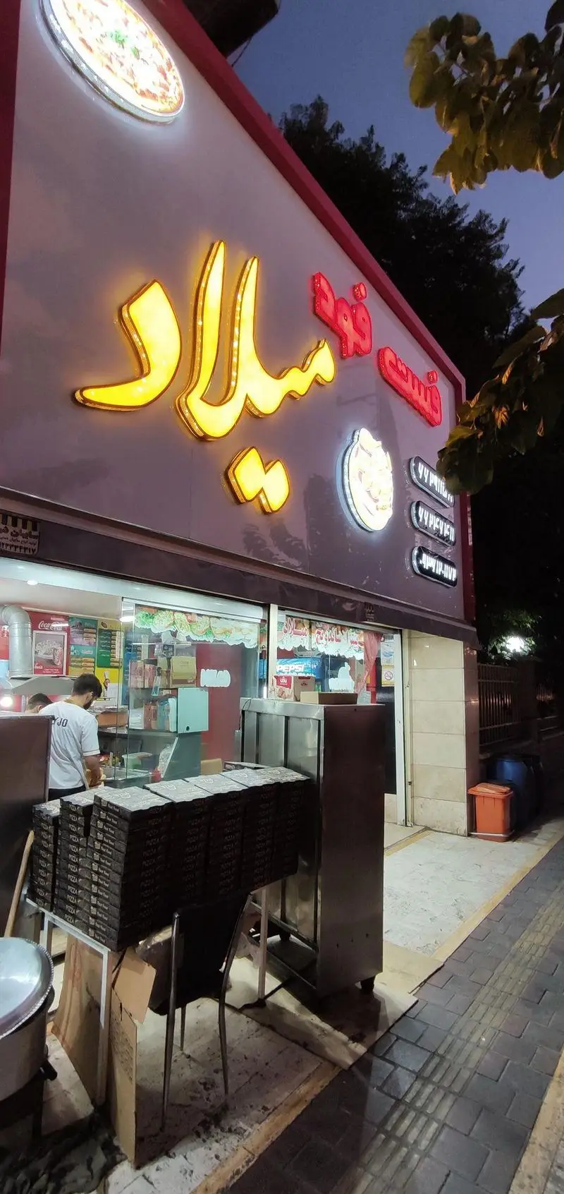 نمای بیرونی فست فود پیتزا میلاد در شب با تابلوهای روشن و جعبه‌های پیتزا آماده تحویل، جذاب برای سفارش آنلاین.