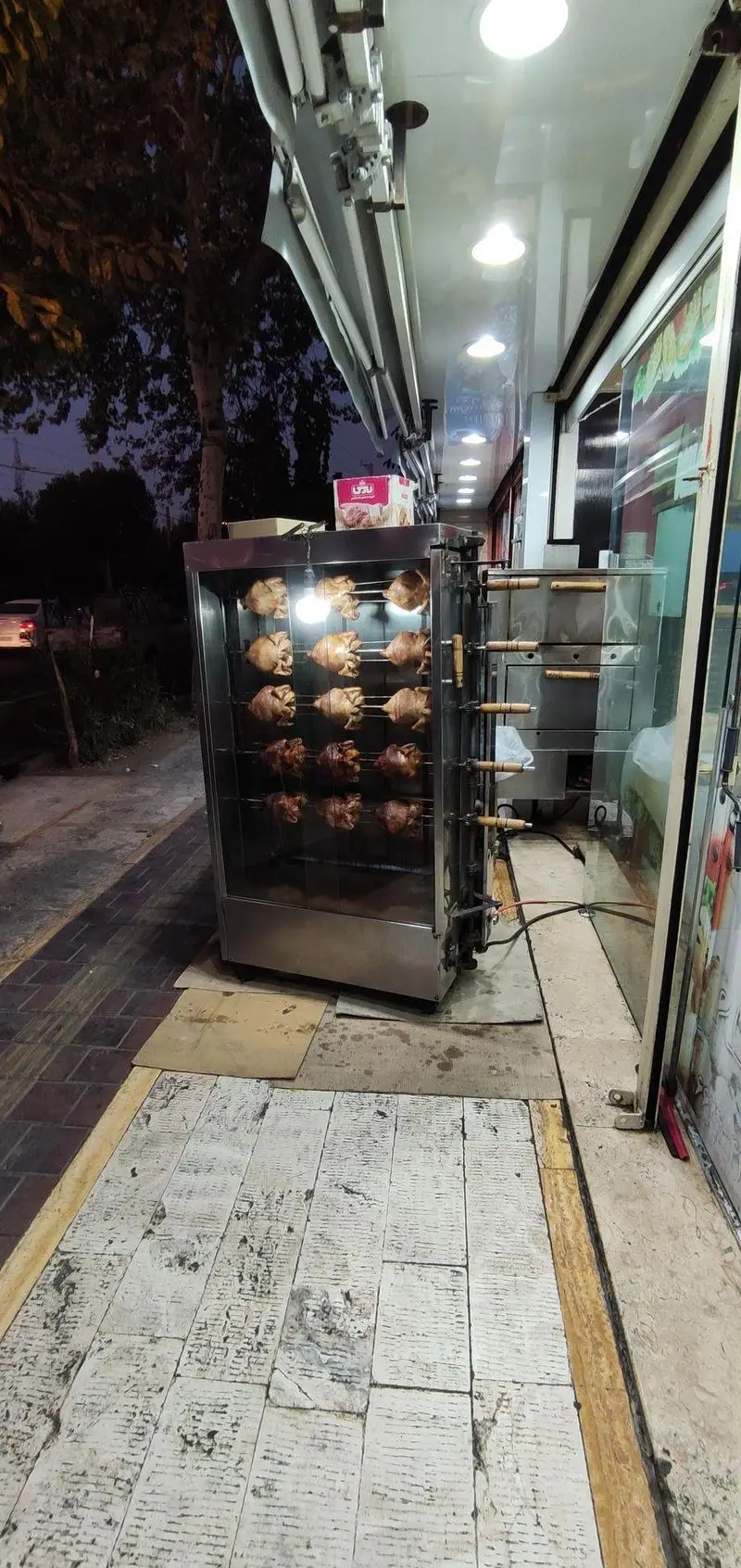 مرغ بریان‌های طلایی پیتزا میلاد، پخته شده در دستگاه جوجه گردان در فضای باز، آماده برای سفارش غذای آماده و فست فود.