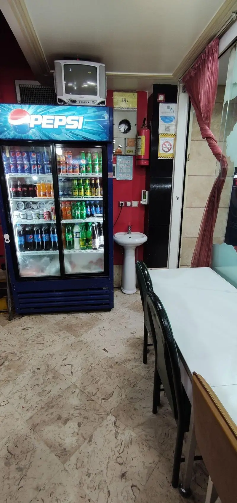 فضای داخلی دنج پیتزا میلاد شامل یخچال پپسی، تلویزیون قدیمی، سینک، کپسول آتش‌نشانی و میز و صندلی‌های تمیز برای مشتریان.