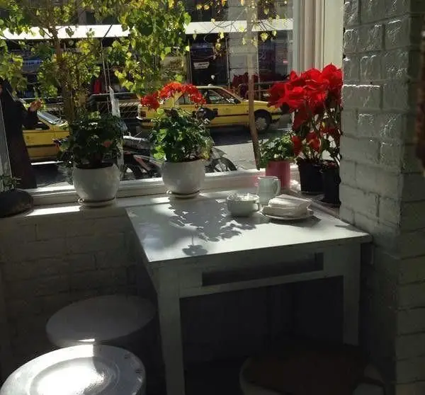 نمایی دنج از کافه قنادی ورتا با میز و صندلی سفید، گلدان‌های گل زیبا کنار پنجره مشرف به خیابان شهر و تاکسی‌های زرد.