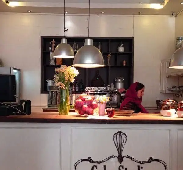 نمای داخلی کافه قنادی ورتا با پیشخوان چوبی، گلدان گل، اپل و پرسنل خانم مشغول کار. محیط گرم و دلنشین شیرینی‌فروشی.