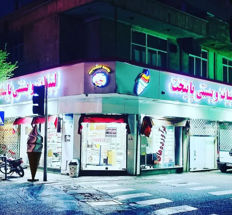 نمای شبانه مغازه لبنیات و بستنی پایتخت با تابلوهای نئونی درخشان، مجسمه بستنی قیفی و چراغ راهنمایی در خیابان
