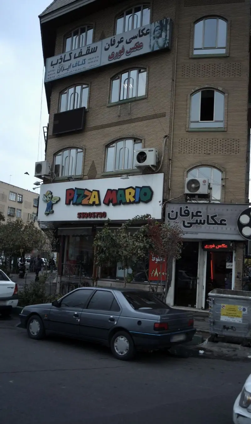 عکس نمای خیابانی پیتزا ماریو، یک فست فود محبوب با تابلوی رنگی "Pizza Mario" و شماره تماس، در کنار یک آتلیه عکاسی.