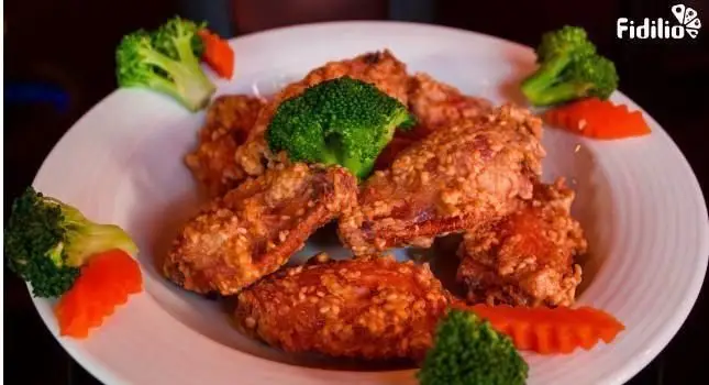 بال مرغ کنجدی ترد و لذیذ، غذای محبوب رستوران چینی اژدهای طلایی، با تزئین بروکلی و هویج تازه برای یک تجربه آسیایی.