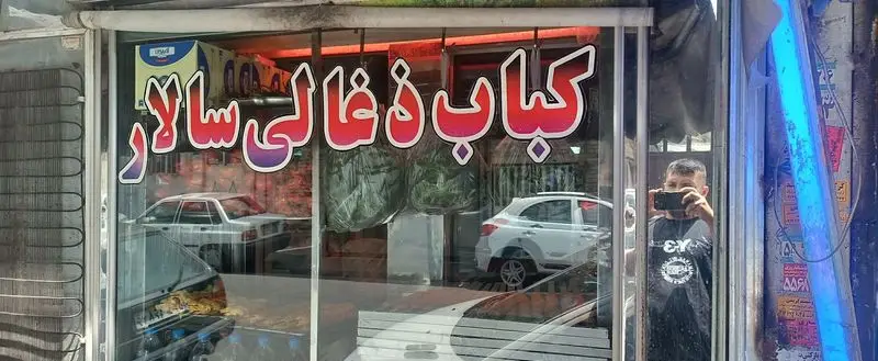 نمای جلوی مغازه کباب ذغالی سالار با تابلوی رنگارنگ و برجسته، شامل انعکاس خودروها، خیابان و شخص عکاس، یک کبابی پرطرفدار.
