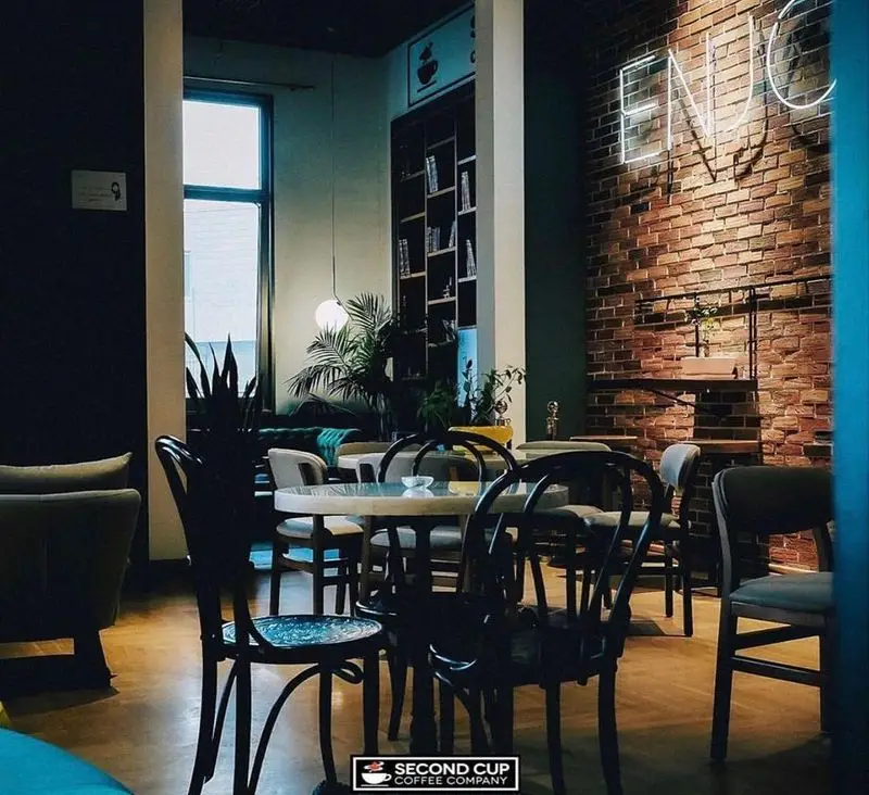 فضای داخلی کافه سکند کاپ با میز و صندلی‌های شیک، دیوار آجری، نئون "ENJOY"، قفسه کتاب و گیاهان سرسبز، محیطی دلنشین.