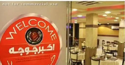 تصویر تابلوی ورودی "اکبر جوجه شعبه چهارراه ولیعصر" همراه با نمای داخلی رستوران، شامل چیدمان میز و صندلی‌ها.