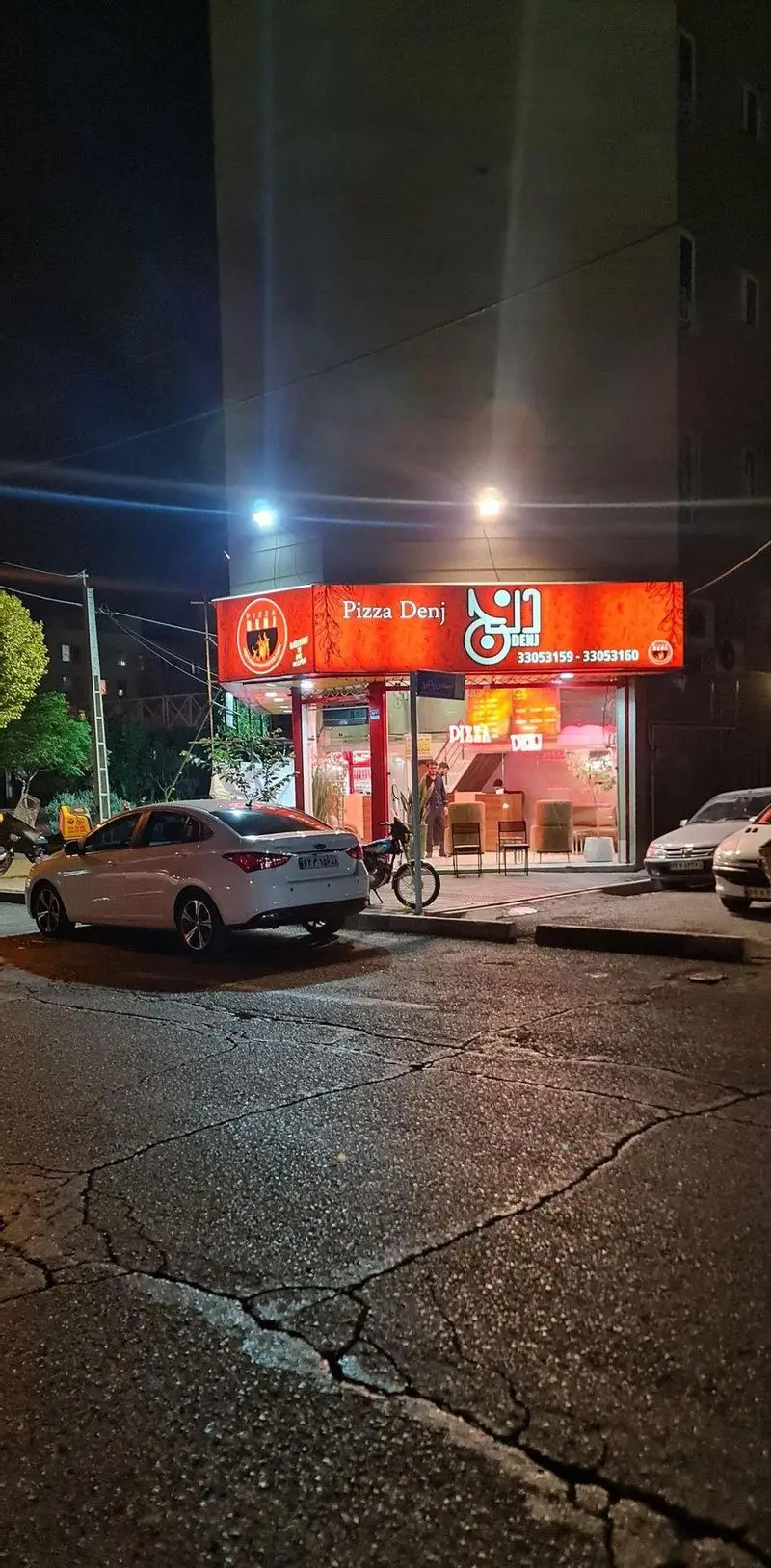 تصویر شبانه از پیتزا دنج، فست فودی مدرن با تابلوی قرمز و چراغ‌های روشن، خودروها در خیابان و نمای کلی شلوغ شهر.