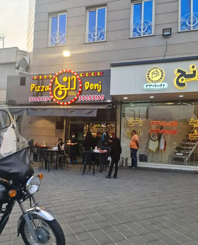 نمای بیرونی رستوران پیتزا دنج در کنار املاک دنج با مشتریان در حال غذا خوردن، تابلوی پیتزا و املاک دنج و شماره تماس قابل مشاهده است.