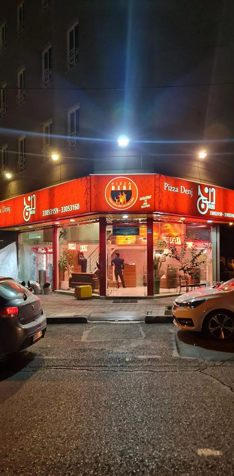 نمای بیرونی و روشن رستوران پیتزا دنج در شب، با تابلوی قرمز و شماره تلفن‌ها، پرسنل داخل و خودروهای پارک شده، مناسب برای سفارش فست فود و تحویل فوری.