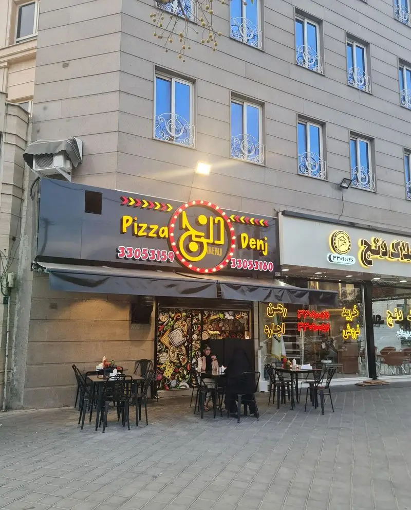 تصویر ورودی رستوران پیتزا دنج با فضای بیرونی، شامل تابلوی روشن پیتزا دنج و شماره‌های تماس 33053159 و 33053160.