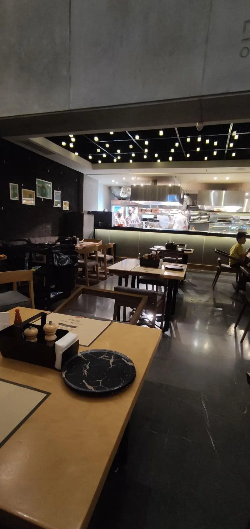 فضای داخلی و دکوراسیون مدرن فست فود دکتر آرین با آشپزخانه باز، میز و صندلی چوبی و کفپوش مشکی، و نورپردازی سقف جذاب.