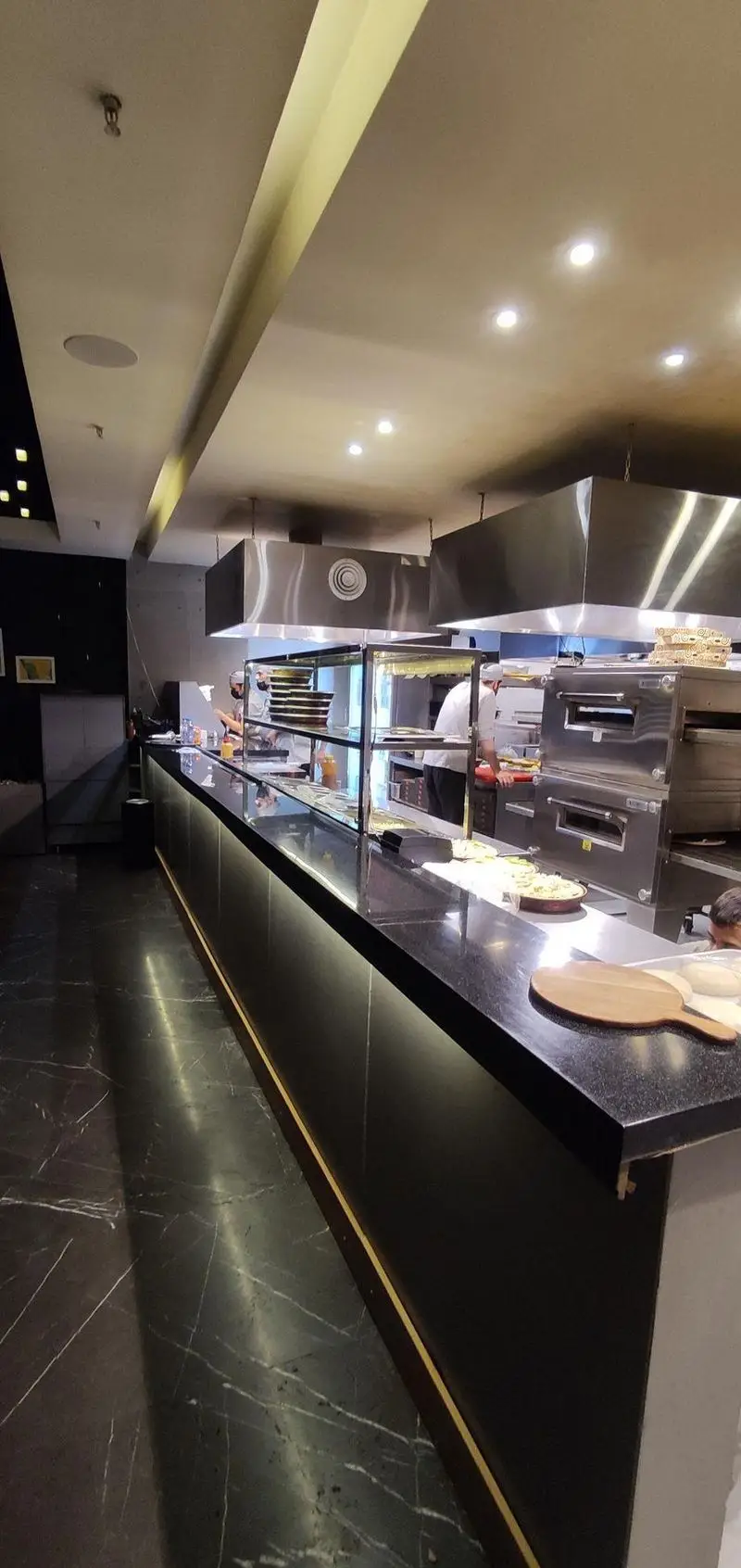 فضای داخلی فست فود دکتر آرین، آشپزخانه باز و مدرن با سرآشپزهای مشغول تهیه پیتزا و غذاهای آماده، کانتر براق و کف مرمری