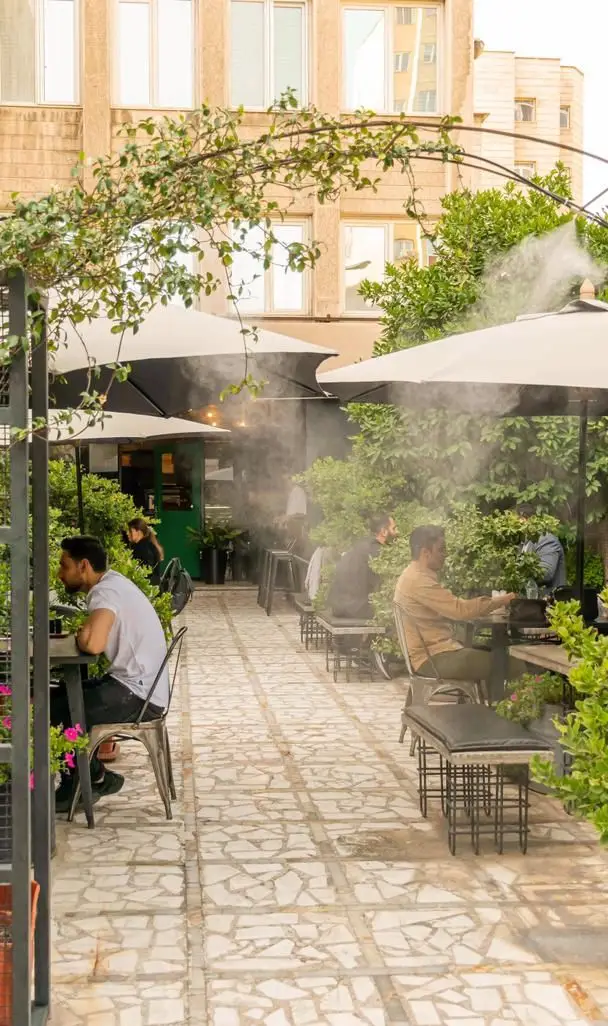نمایی دلنشین از فضای باز کافه رام بیسترو، مشتریان در حال لذت از غذا و نوشیدنی زیر سایه‌بان‌های مه‌پاش‌دار خنک.