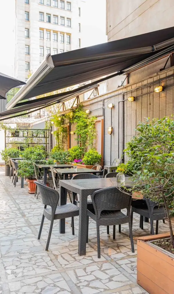 نمایی از فضای باز دنج و سرسبز کافه رام بیسترو با میز و صندلی‌های مشکی حصیری، آماده پذیرایی با انواع غذا و نوشیدنی‌های خوشمزه.