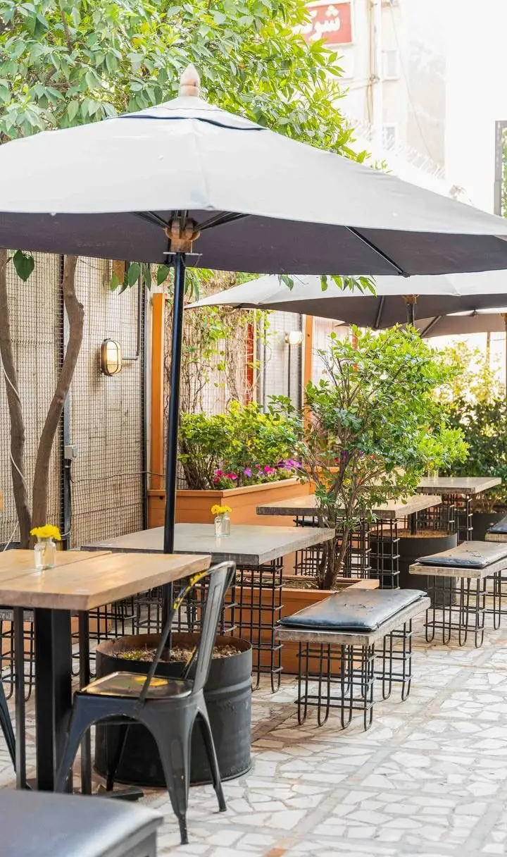 تصویر فضای باز کافه رام بیسترو با میز و صندلی‌های راحت، چترهای بزرگ و محیطی پر از گیاهان سبز، مناسب برای دورهمی و صرف انواع غذا و نوشیدنی.