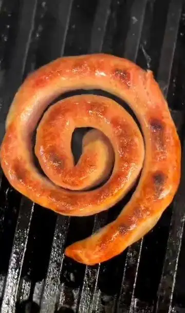 سوسیس حلزونی آبدار و طلایی رنگ در حال گریل شدن روی توری داغ، نمادی از غذاهای وسوسه‌انگیز کافه رام بیسترو با نوشیدنی‌های متنوع