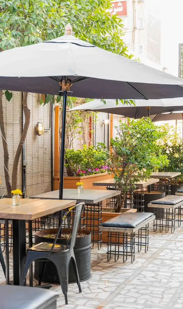 فضای باز کافه رام بیسترو با میز و صندلی‌های چوبی و فلزی، چترهای سایبان بزرگ، و گیاهان سرسبز، محیطی دلنشین برای صرف انواع غذا و نوشیدنی.