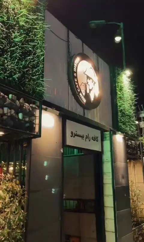 تصویری از نمای بیرونی کافه رام بیسترو در شب، با تابلوی نورانی Rum Cafe & Bistro و ورودی سرسبز برای انواع غذا و نوشیدنی.