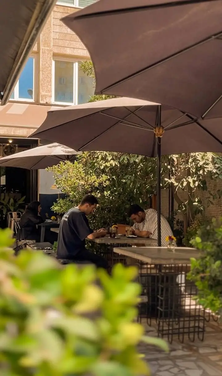 مشتریان در فضای باز کافه رام بیسترو تهران زیر چترها، از انواع غذا و نوشیدنی در محیطی سرسبز و دلنشین لذت می‌برند.