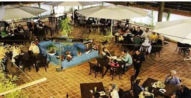 نمای هوایی از فضای دلنشین و باز پیتزایی طلوع در شب، مشتریان شاد در حال غذا خوردن زیر چترهای بزرگ کنار حوض آب.