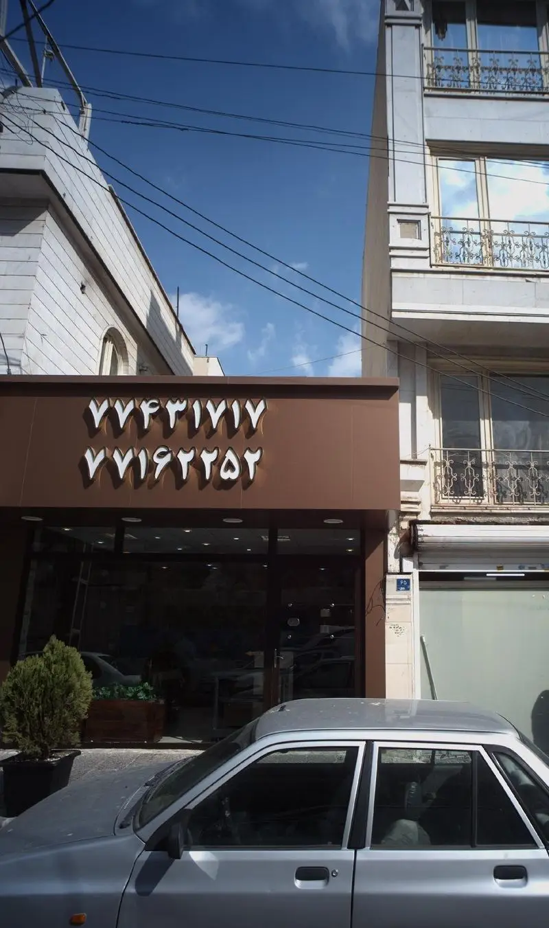 نمای خارجی رستوران صلوات با شماره تلفن‌های ۷۷۴۳۱۷۱۷ و ۷۷۱۶۲۲۵۲، یک صحنه شهری در روز آفتابی با ساختمان‌های اطراف و یک ماشین پارک شده در جلو.