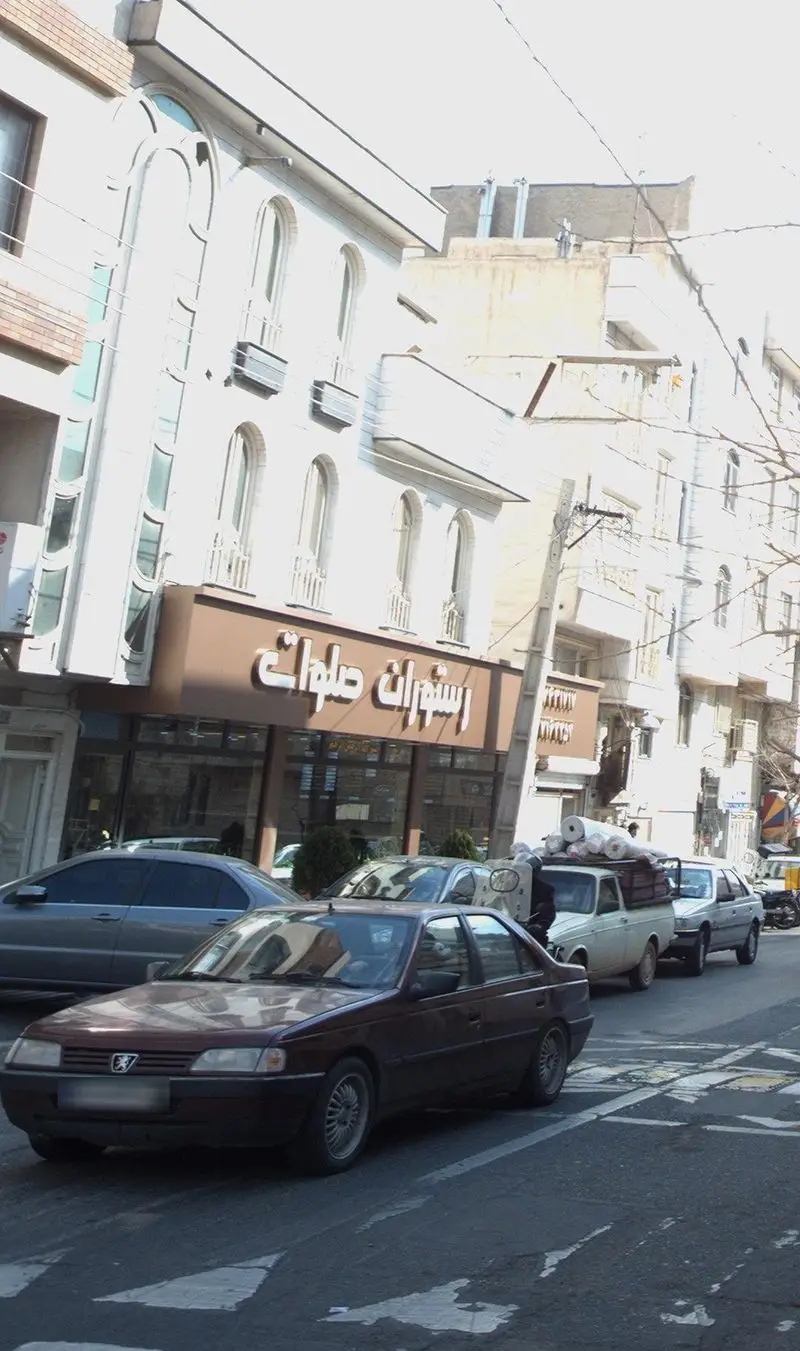 تصویری از نمای بیرونی رستوران صلوات در یک روز آفتابی، شامل تابلوی بزرگ قهوه‌ای و ساختمان سفید با طراحی سنتی و خودروها در جلو.