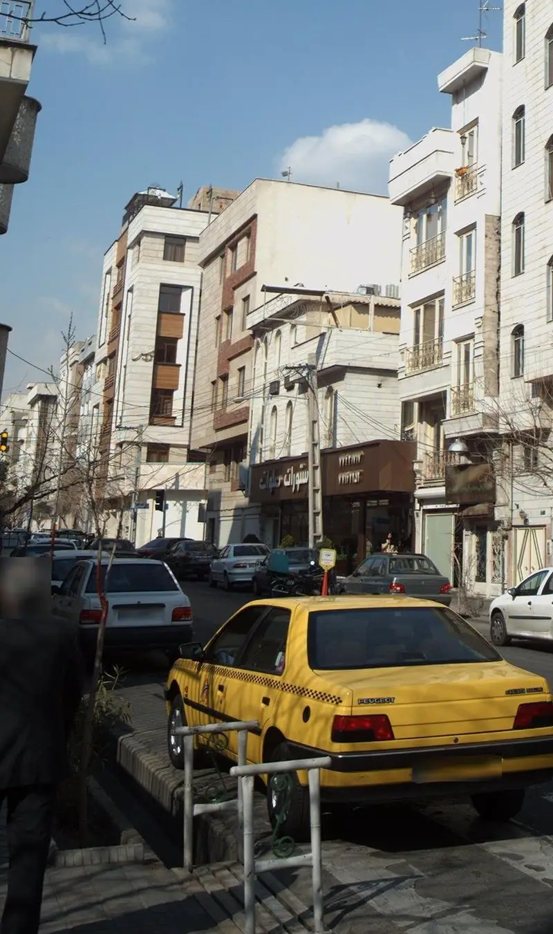 رستوران صلوات در خیابان شلوغی با ساختمان‌های بلند و خودروهای پارک‌شده، به همراه تاکسی زرد رنگ در پیش‌زمینه. فضای شهری ایران.