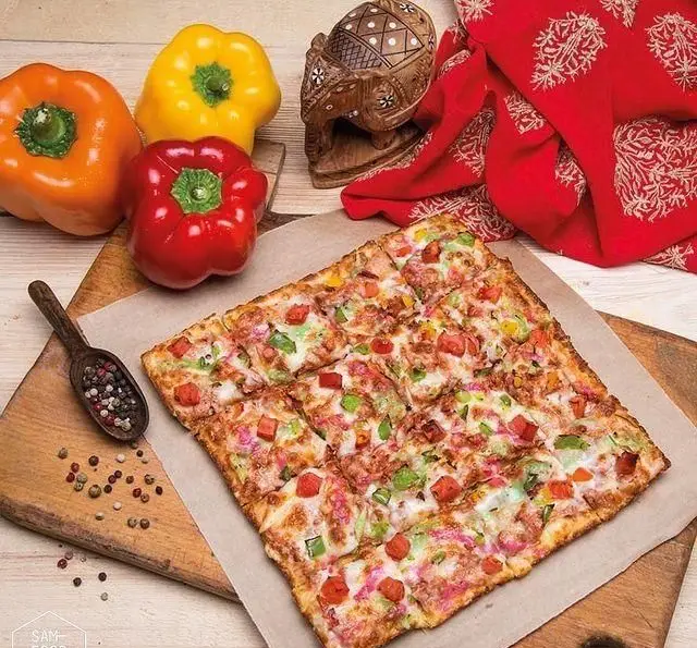 پیتزا مربعی خاص فودهال سام، تازه پخته شده با فلفل دلمه ای رنگارنگ، ادویه جات معطر، روی تخته چوبی، منتظر سفارش شماست.