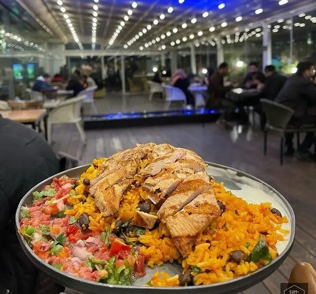یک بشقاب بزرگ حاوی سینه مرغ گریل، برنج زرد با حبوبات و سالسا در فودهال سام. فضای داخلی مدرن و نورپردازی زیبای رستوران.