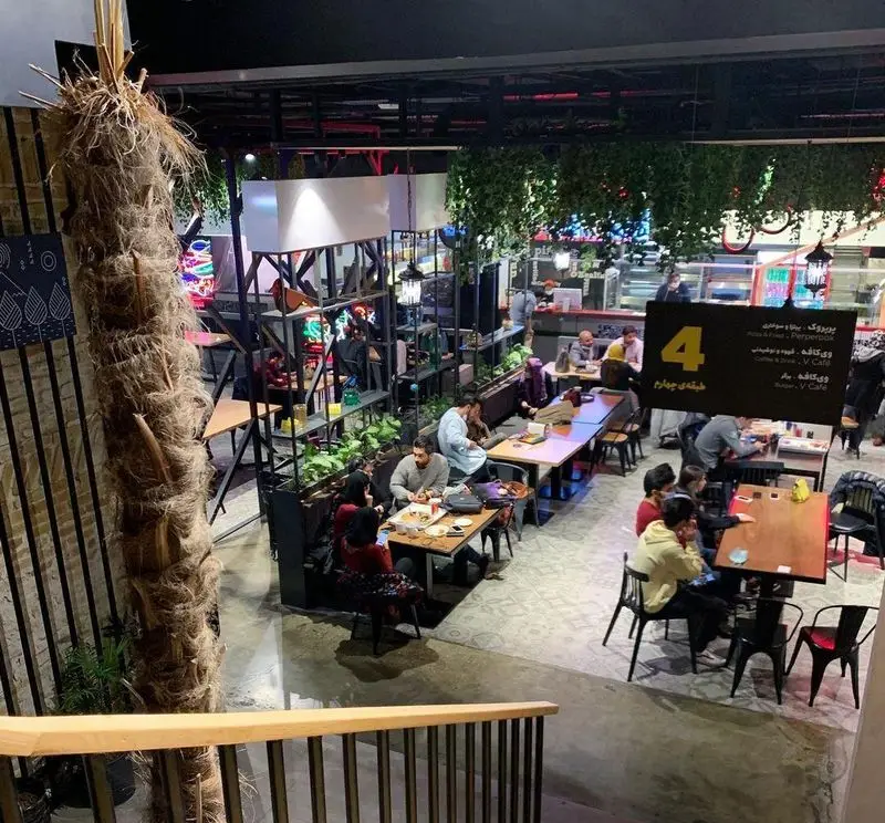 نمای داخلی شیک و مدرن فودهال سام با مشتریانی در حال صرف غذا، میز و صندلی‌های چوبی و فلزی، گیاهان آویز و نورپردازی جذاب.