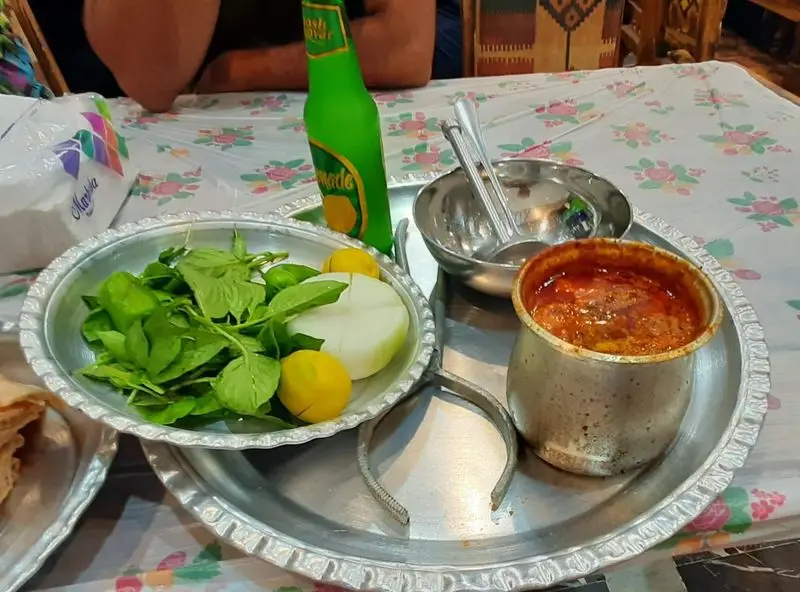 وعده غذایی دیزی اصیل و خوش طعم در رستوران دیزی علی بابا؛ سرو شده با سبزی تازه، پیاز، لیمو و نوشیدنی روی سینی فلزی.