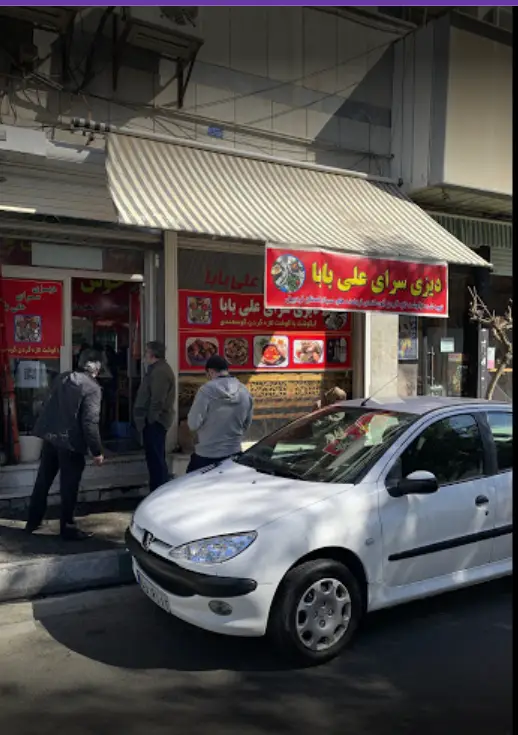 نمای بیرونی دیزی سرای علی بابا، رستوران محبوب غذای سنتی دیزی با تابلوهای قرمز، مشتریان و پژو ۲۰۶ سفید در خیابان شهر.