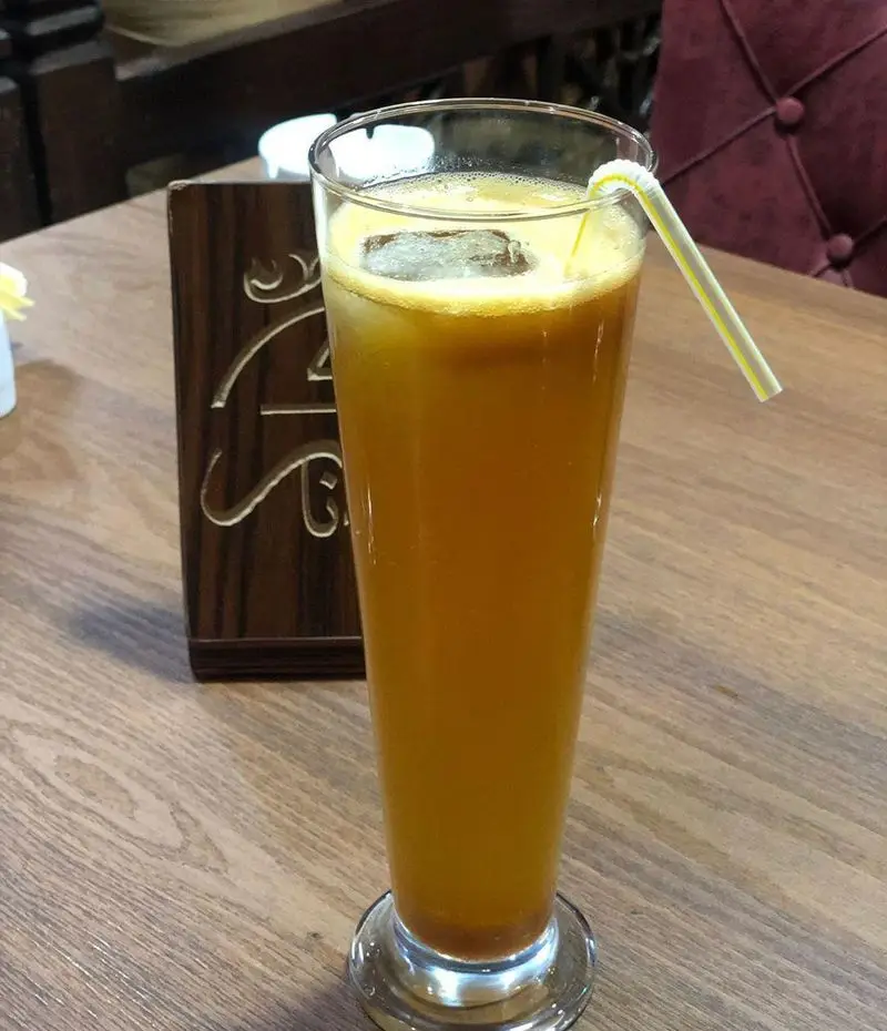 تصویری از یک نوشیدنی گوارا و خنک با یخ و نی در لیوانی بلند روی میز چوبی، در فضای دلنشین سفره خانه سنتی دیزی نار.