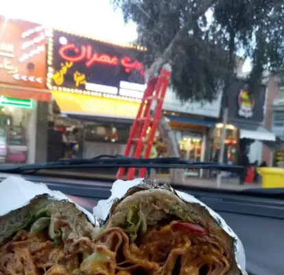 ساندویچ‌های بزرگ و خوشمزه فست فود بمب مهران داخل خودرو، با تابلوی نئون روشن رستوران در پس‌زمینه خیابان شلوغ.