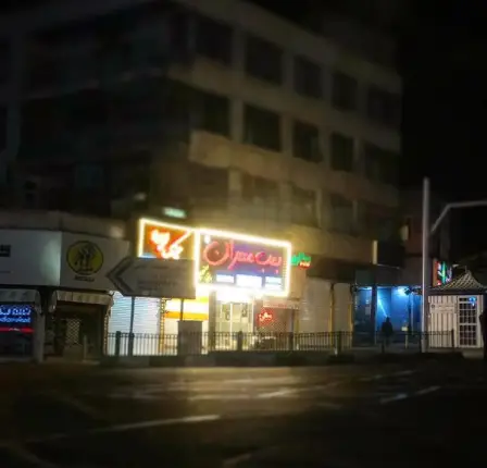 فست فود بمب مهران با تابلوی نئون درخشان در شب، آماده سرو انواع غذاهای خوشمزه و ساندویچ های هیجان انگیز در محیطی دلپذیر و مدرن.