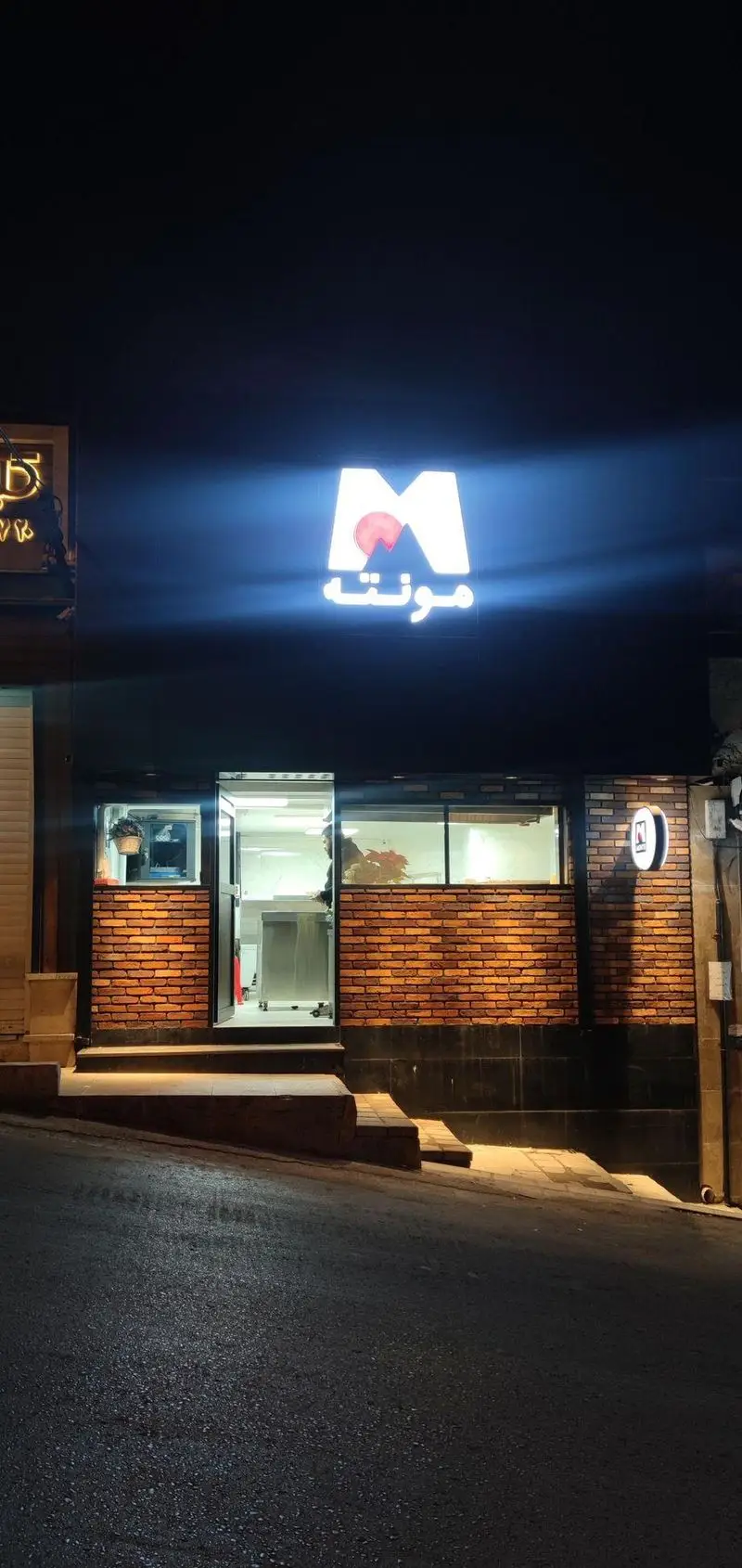 نمای بیرونی و تابلوی روشن آشپزخانه مونته در شب، با لوگوی M کوهستان و نوشته مونته فارسی، ورودی و پنجره‌های آشپزخانه آجری.