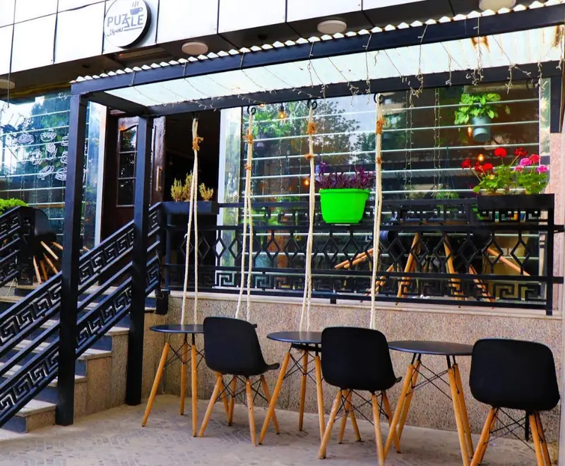 نمای بیرونی کافه پازل با میز و صندلی‌های چوبی و مشکی مدرن، نرده‌های طرح‌دار، گل و گیاه آویزان و چراغ‌های تزئینی برای مشتریان.