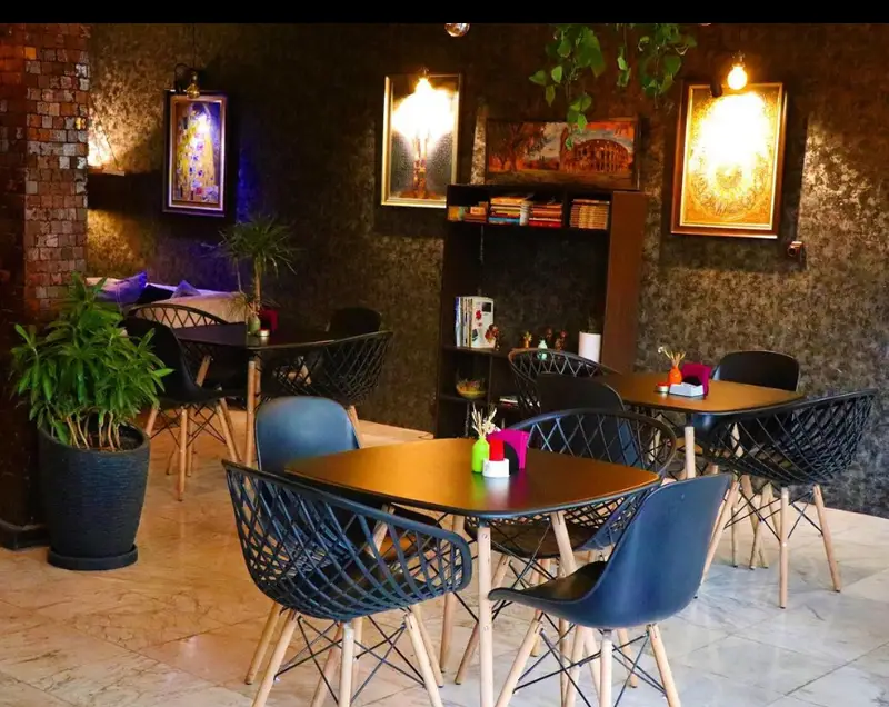 فضای داخلی کافه پازل با دکوراسیون شیک، میز و صندلی‌های مدرن، گلدان‌های سبز، دیوارهای طرح‌دار و تابلوهای هنری در محیطی دنج و آرام.