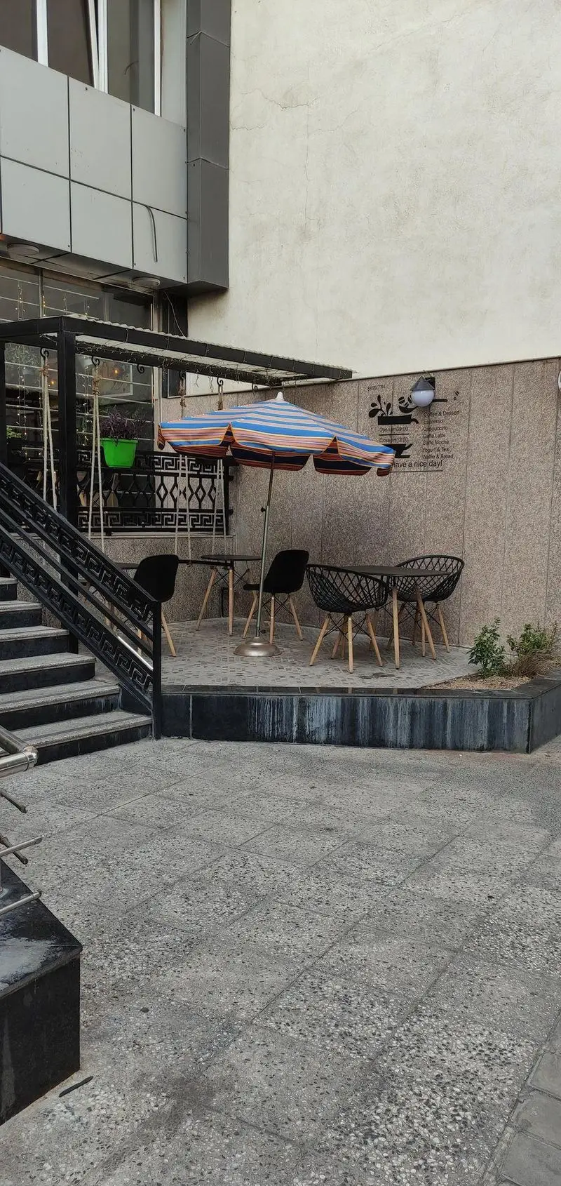 نمای بیرونی کافه پازل، شامل فضای باز با میز و صندلی‌های مدرن، چتر راه راه، پله‌ها و منوی نوشیدنی‌ها. محیطی آرام برای قهوه.