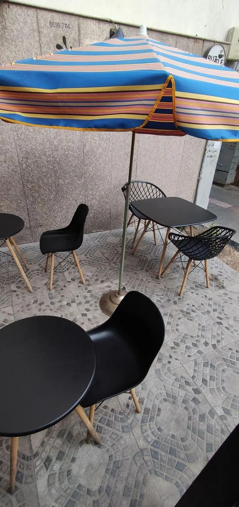 کافه پازل: فضای بیرونی با میز و صندلی‌های مشکی مدرن، چتر بزرگ راه راه آبی و نارنجی، محیطی دلپذیر برای مشتریان.