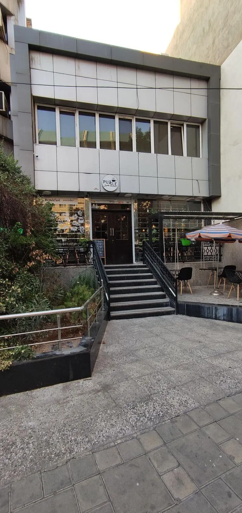 کافه پازل (PUZZLE Cafe) با ورودی پلکانی، فضای نشیمن روباز، چتر رنگی و تابلوی "Breakfast everyday" در محیطی دنج و مدرن.